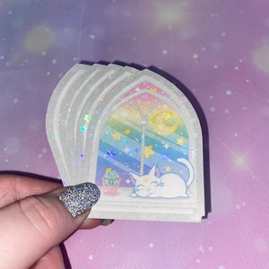 May include: A set of four iridescent stickers featuring a white cat sleeping in a window with a rainbow and stars. The stickers are on a white background.