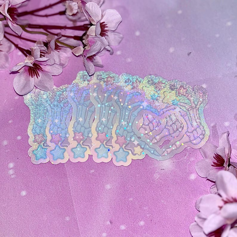 May include: A set of five iridescent holographic stickers featuring a skeleton hand design with stars. The stickers are on a pink and purple background.