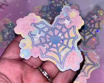 May include: A pastel pink, blue, and yellow iridescent sticker with a heart-shaped spiderweb design. The spiderweb has a blue heart in the center and is surrounded by pink cherry blossoms and white stars.
