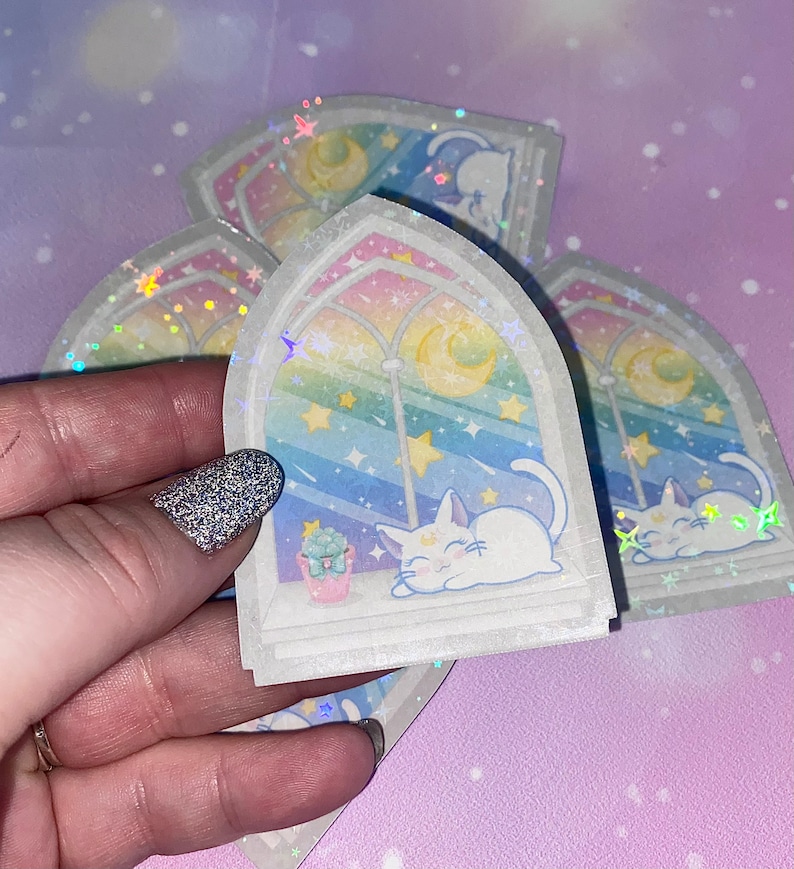 May include: A set of five iridescent stickers featuring a white cat sleeping in a window with a rainbow, stars, and a crescent moon. The stickers are on a pink background.