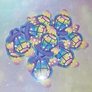 May include: A set of colorful holographic stickers featuring a rainbow gem design with stars. The stickers are on a light blue background with a galaxy pattern.