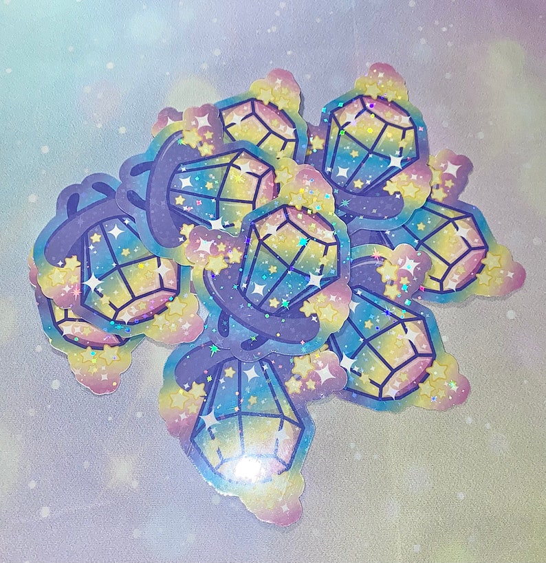 May include: A set of colorful holographic stickers featuring a rainbow gem design with stars. The stickers are on a light blue background with a galaxy pattern.