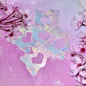 May include: A set of iridescent holographic stickers featuring skeleton hands forming hearts with stars and glitter. The stickers are on a pink and purple background.