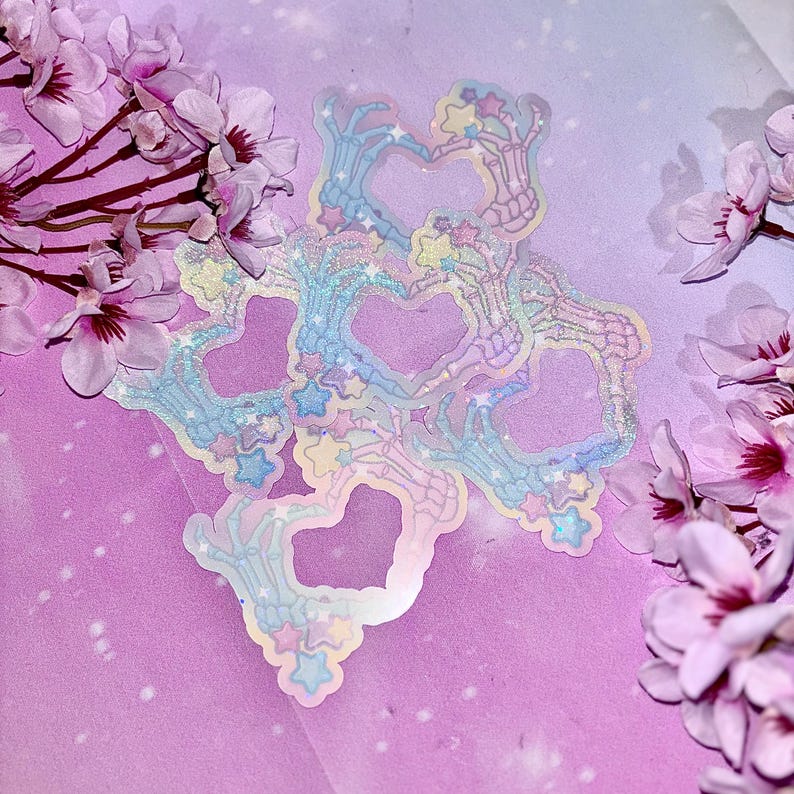 May include: A set of iridescent holographic stickers featuring skeleton hands forming hearts with stars and glitter. The stickers are on a pink and purple background.