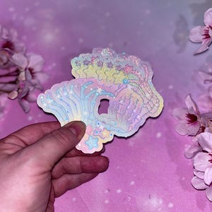 May include: A holographic sticker featuring a rainbow skeleton hand with stars and clouds. The sticker is iridescent and has a glittery finish.