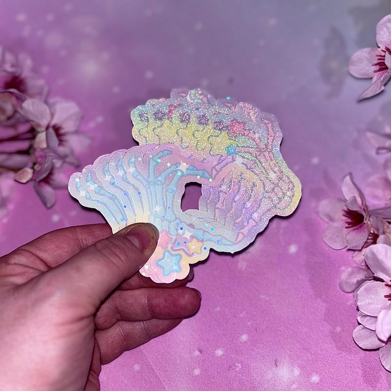 May include: A holographic sticker featuring a rainbow skeleton hand with stars and clouds. The sticker is iridescent and has a glittery finish.