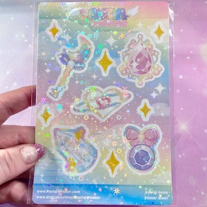May include: A sheet of iridescent holographic stickers featuring kawaii gems, stars, and other celestial designs. The sticker sheet is packaged in a clear plastic sleeve and has the text "Kawaii Gems Sticker Sheet" printed on the bottom right corner.