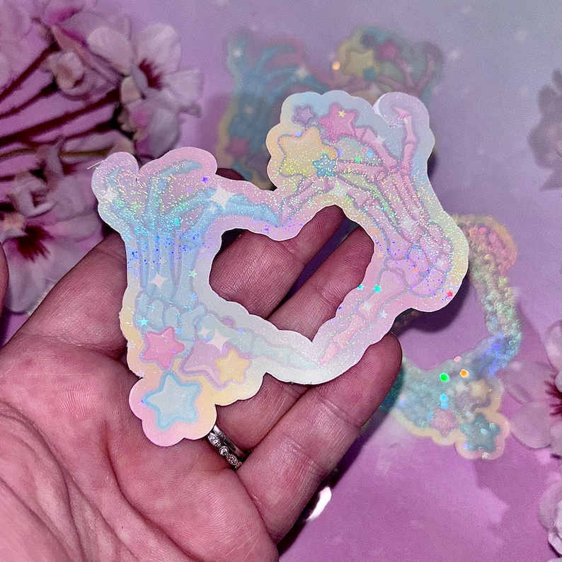 May include: Two iridescent skeleton hands forming a heart shape with a glittery, pastel background. The hands are outlined in white and have a rainbow glitter effect. The background is a soft pink with a subtle shimmer.