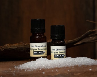 Op de afbeelding: Twee kleine bruine glazen flesjes met zwarte dopjes en witte etiketten. De etiketten zeggen "Your Choice here 100% pure Essential Oil" en "HALKAB Organics". De flesjes staan op een houten oppervlak met witte suikerkristallen.