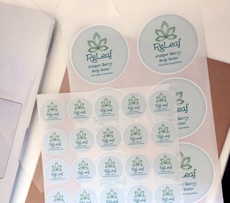 May include: A collection of round stickers with a light blue background and a green leaf design. The text "ReLeaf Juniper Berry Body Butter 100% Natural Ingredients" is printed on the stickers. Some stickers are on a clear sheet.