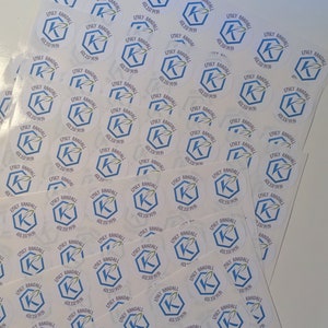 May include: A collection of white, circular stickers featuring the name "Emily Randall" and a phone number. Each sticker has a blue hexagon and a stylized "K" with a green leaf design. The stickers are arranged in stacks.
