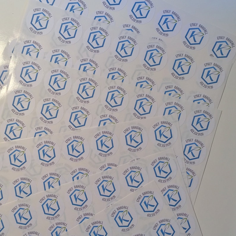 May include: A collection of white, circular stickers featuring the name "Emily Randall" and a phone number. Each sticker has a blue hexagon and a stylized "K" with a green leaf design. The stickers are arranged in stacks.