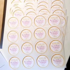May include: A stack of round stickers with gold borders and text. The stickers say "THANK YOU FOR COMING! Ivie & Ekan's 1ST BIRTHDAY 18/08/2018" in pink and gold. The stickers are on a white surface.