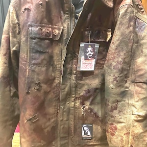 May include: Brown and tan jacket with a distressed, blood-splattered design. Features a corduroy collar, multiple pockets, and a zip-up front. Includes patches and tags with graphic imagery. A unique, themed outerwear piece.
