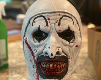 May include: A white clown mask with black eye markings, a black mouth, and a small black top hat. The mask has red blood-like streaks and splatters. The clown's teeth are visible and stained. The mask is a Halloween costume.