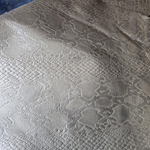 Könnte beinhalten: Nahaufnahme einer hellgrauen Kunstlederplatte mit Schlangenmuster. Das geprägte Design weist Schuppen und unregelmäßige Formen auf, wodurch eine strukturierte Oberfläche entsteht. Das Material ist gefaltet und zeigt die Unterseite.