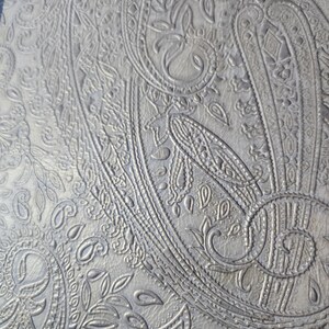 May include: Close-up of a silver-gray paisley pattern. The design features intricate, embossed details of swirling floral and teardrop shapes. The texture appears to be leather or a similar material, with a subtle sheen.