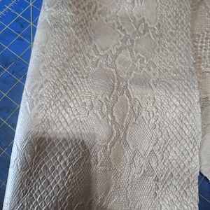 Pode incluir: Close-up de um pedaço de tecido de pele de cobra sintética. O tecido tem uma superfície texturizada com um padrão de escamas em tons de cinza e branco sujo. O material está dobrado, revelando o verso.