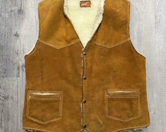 May include: A brown suede vest with a cream-colored, fuzzy lining and trim. The vest has two front pockets, a button-up closure, and decorative stitching. The vest is a vintage style.