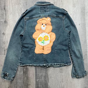 May include: A blue denim jacket with a Care Bear patch on the back. The Care Bear is peach colored with a yellow flower and two yellow smiling suns on its chest.