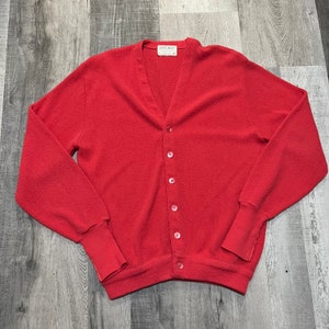 May include: A red cardigan sweater with a v-neck and button closure. The sweater has long sleeves and ribbed cuffs. The label on the sweater reads "Geoffrey Beene".