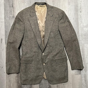 May include: A brown tweed blazer featuring a textured speckled pattern, a notched lapel, two front buttons, and two flap pockets. The interior is lined with a smooth, light beige fabric, and the garment includes a single chest welt pocket.
