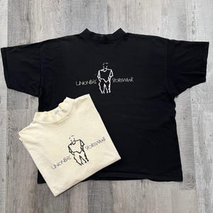 May include: Two t-shirts, one black and one cream-colored, with a mock neck design. Each shirt features a white graphic of a person and the text "UNIONBAY SPORTSWEAR". The shirts are laid flat on a wooden surface.