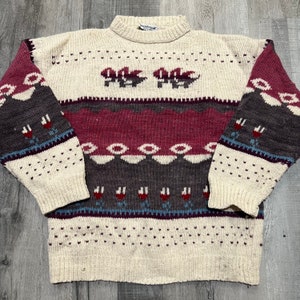 May include: A cream-colored knitted sweater with a multi-colored geometric pattern. The pattern includes shades of pink, brown, blue, and red. The sweater has a crew neck and long sleeves.