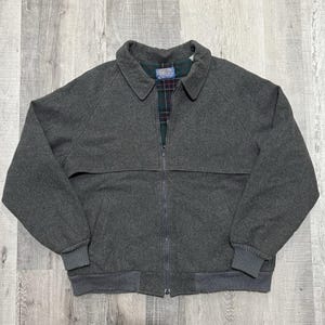 May include: A charcoal gray wool jacket with a full zipper closure. The jacket features a collared neckline, ribbed cuffs, and a ribbed hem. The interior lining is a plaid pattern of green, red, and black. A classic outerwear piece.