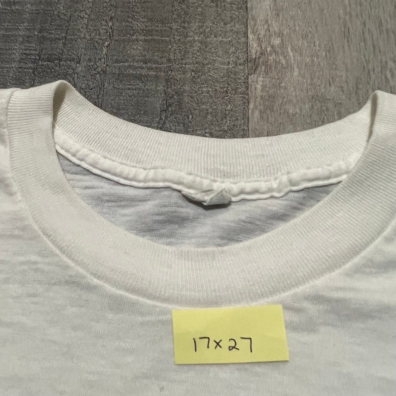 Puede incluir: Camiseta blanca de cuello redondo con una etiqueta que dice "17 x 27".