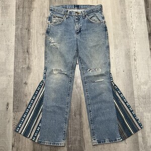 May include: Light blue denim jeans with distressed details and flared bottoms. The jeans feature decorative fabric panels with a geometric pattern in shades of blue, white, and brown along the outer seams.