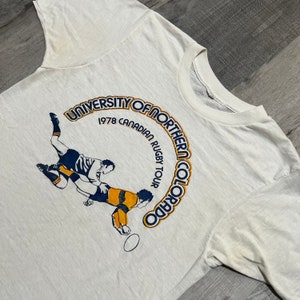 Puede incluir: Camiseta blanca con un gr&aacute;fico azul y amarillo de dos jugadores de rugby en acci&oacute;n. El gr&aacute;fico est&aacute; rodeado por un c&iacute;rculo con el texto "University of Northern Colorado 1978 Canadian Rugby Tour".