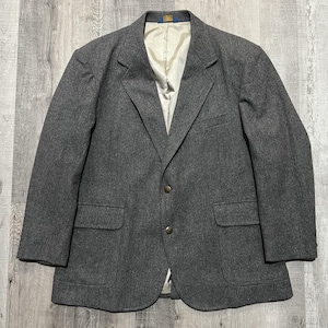 May include: A charcoal gray wool blazer featuring a notched lapel, two front buttons, and two flap pockets. The interior is lined with a smooth, light cream-colored fabric. The garment has long sleeves and a structured, tailored silhouette.