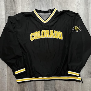 May include: A black long-sleeve pullover windbreaker featuring the word COLORADO in yellow block letters across the chest. The V-neck collar, cuffs, and hem have white and yellow stripes. A yellow buffalo logo is on the upper left sleeve.
