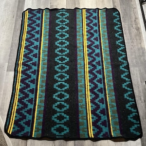 May include: A rectangular blanket with a black border. The blanket features a repeating pattern of teal, purple, and yellow vertical stripes against a dark gray background. The design includes geometric shapes and zigzags.