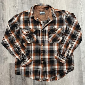 May include: A brown, black, and white plaid flannel shirt with two chest pockets and button closures. The shirt has a brown quilted lining and a collar. The shirt is laid flat on a wooden surface.