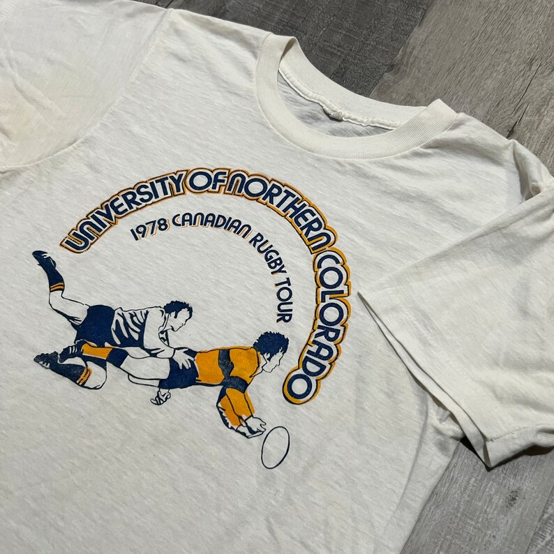 Puede incluir: Camiseta blanca con un gr&aacute;fico azul y amarillo de dos jugadores de rugby. El gr&aacute;fico est&aacute; en un c&iacute;rculo con el texto "University of Northern Colorado 1978 Canadian Rugby Tour" alrededor.