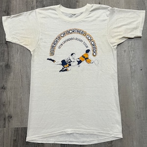 Puede incluir: Camiseta blanca con un gr&aacute;fico azul y amarillo de dos jugadores de rugby en acci&oacute;n. El gr&aacute;fico est&aacute; rodeado por un c&iacute;rculo azul con el texto "University of Northern Colorado 1978 Canadian Rugby Tour".