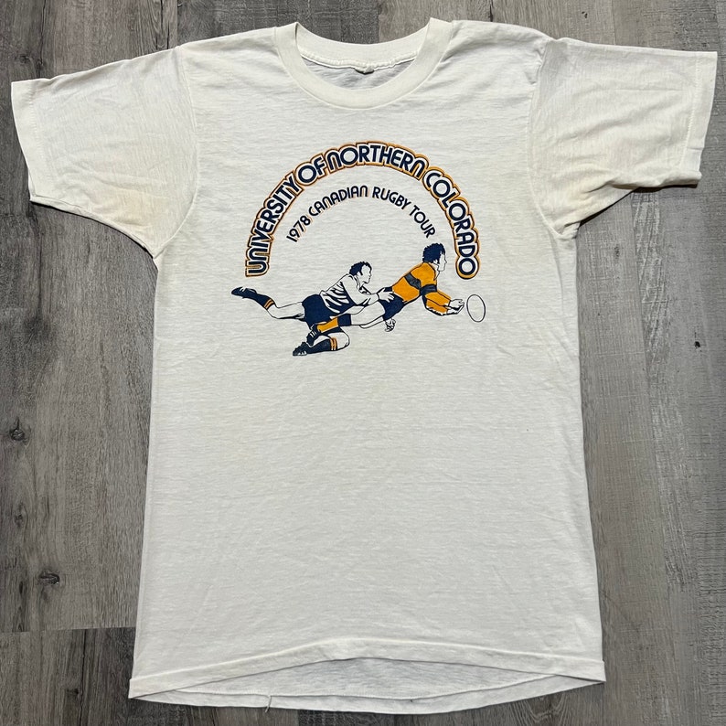 Puede incluir: Camiseta blanca con un gr&aacute;fico azul y amarillo de dos jugadores de rugby en acci&oacute;n. El gr&aacute;fico est&aacute; rodeado por un c&iacute;rculo azul con el texto "University of Northern Colorado 1978 Canadian Rugby Tour".