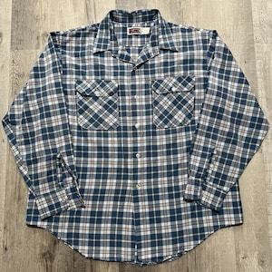 May include: A long-sleeved, button-down shirt with a plaid pattern in shades of blue, white, and burgundy. The shirt features two chest pockets and a classic collar. The fabric appears to be a medium-weight material.
