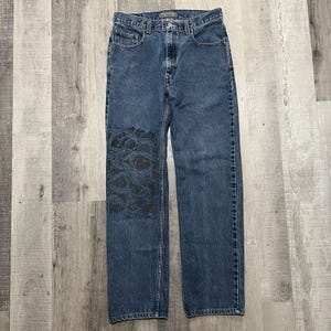 May include: Blue denim jeans with a straight leg cut. The jeans feature a black graphic design of multiple eyes on the left leg. The jeans have a classic five-pocket design and a button and zipper closure.