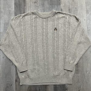 May include: A light gray cable knit sweater with a crew neck and long sleeves. The sweater features vertical cable knit detailing and a small embroidered emblem on the chest. The overall style is casual and comfortable.