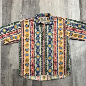 May include: Short-sleeved button-down shirt with a colorful geometric pattern. The shirt features vertical stripes in shades of blue, yellow, red, and green, with circular and star-like designs. The collar and sleeves have a similar patterned design.