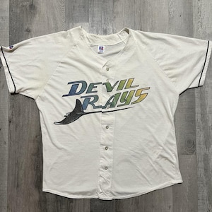 May include: A vintage white baseball jersey with the text "DEVIL RAYS" in blue and yellow. The jersey has a button-up front, black trim on the sleeves, and a logo of a stingray. The jersey is made of a soft, worn material.