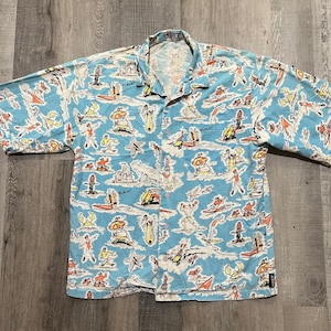 May include: A blue short-sleeved button-up shirt with a cartoon print of people surfing and other beach activities. The shirt has a white background with blue and orange accents. The text "SLAM!" appears multiple times on the shirt.