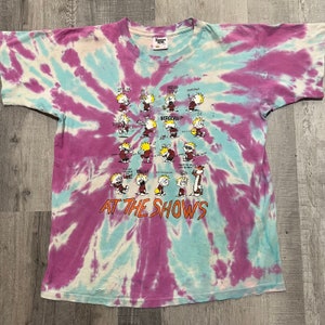May include: A tie-dye t-shirt with a cartoon illustration of Calvin from the comic strip Calvin and Hobbes. The shirt features a variety of scenes of Calvin at a concert, with the text "At The Shows" printed below the illustration.