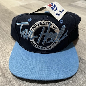 May include: A navy blue and light blue baseball cap with the text "Tar Heels" embroidered on the front. The cap has a flat brim and a tag that reads "The Game". The cap is a classic style.