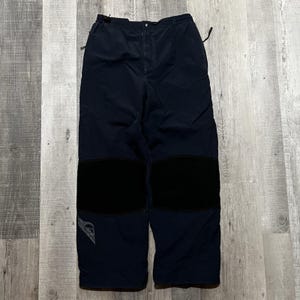 May include: Dark navy blue pants featuring a button and zipper fly closure, an adjustable waistband with side straps, and large black reinforced patches on the knees. A grey logo graphic is visible on the lower right leg of the garment.