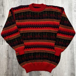 May include: A long-sleeve crew neck sweater featuring a repeating pattern of llamas and geometric Greek key motifs. The knit garment displays horizontal bands of vibrant red, deep black, and dark purple, with ribbed cuffs and a ribbed neckline.