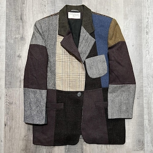May include: A patchwork blazer featuring a variety of fabric textures and patterns, including herringbone, plaid, and solid panels in shades of brown, gray, blue, and tan. The garment has a notched lapel, a single front button, and one chest pocket.